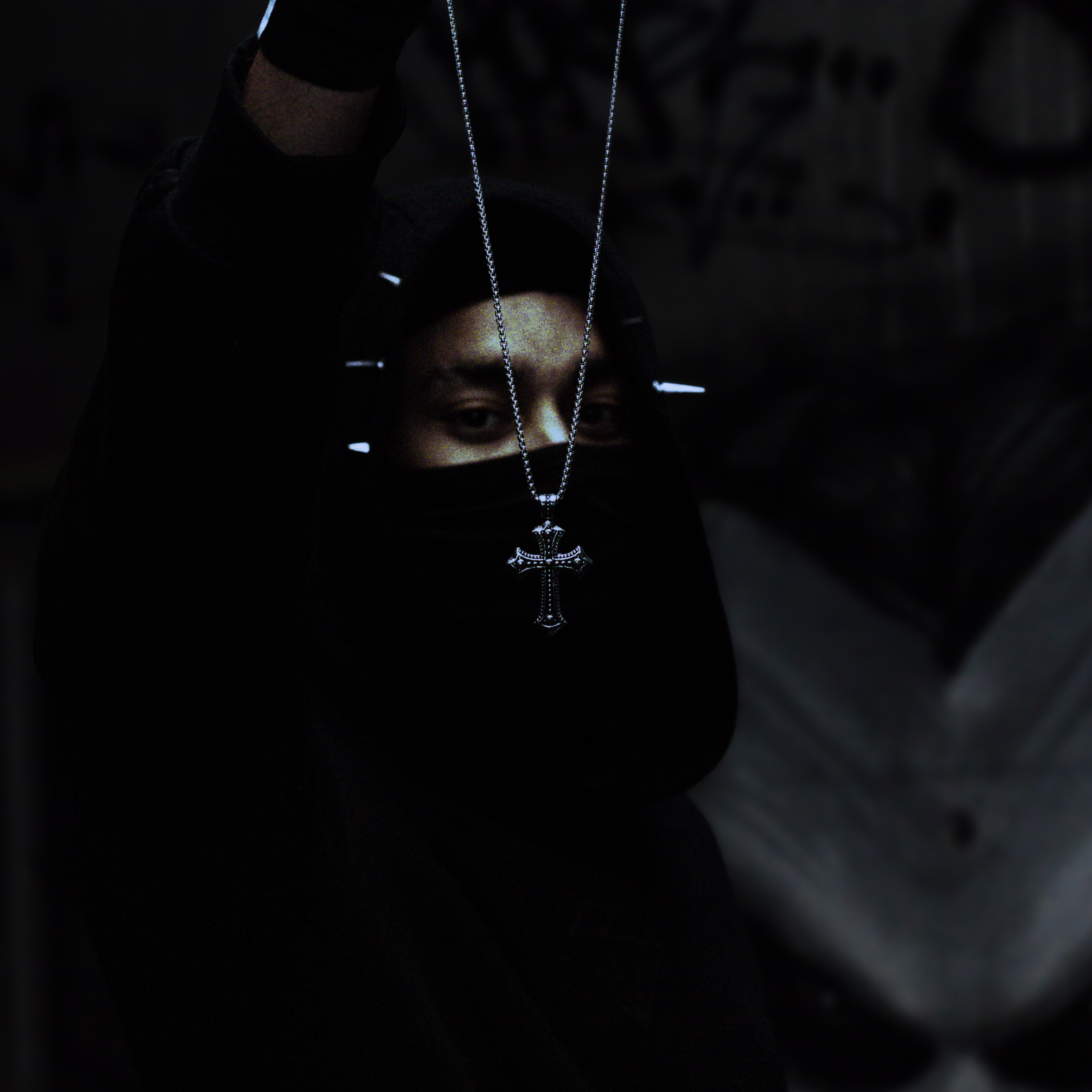 Person wearing a dark hoodie with a necklace featuring a cross pendant in a dimly lit setting.